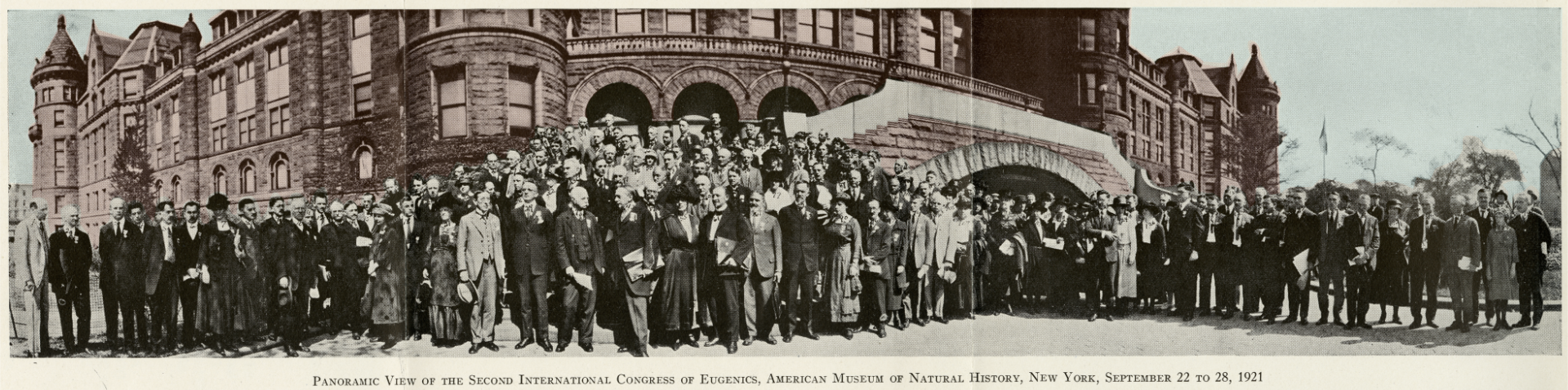 A partially colorized panoramic photo of participants of the Second International Congress of Eugenics posing in front of the American Museum of Natural History building. In the picture, the building is colorized in light brown shade and the participants are in black and white. There are 151 people in the photo whom are mostly men with some women also visible. All participants are well-dressed in the fashion of that era. The caption of the photo reads “Panramic View of the Second International Congress of Eugenics, American Museum of Natural History, New York, September 22 to 28, 2921”.